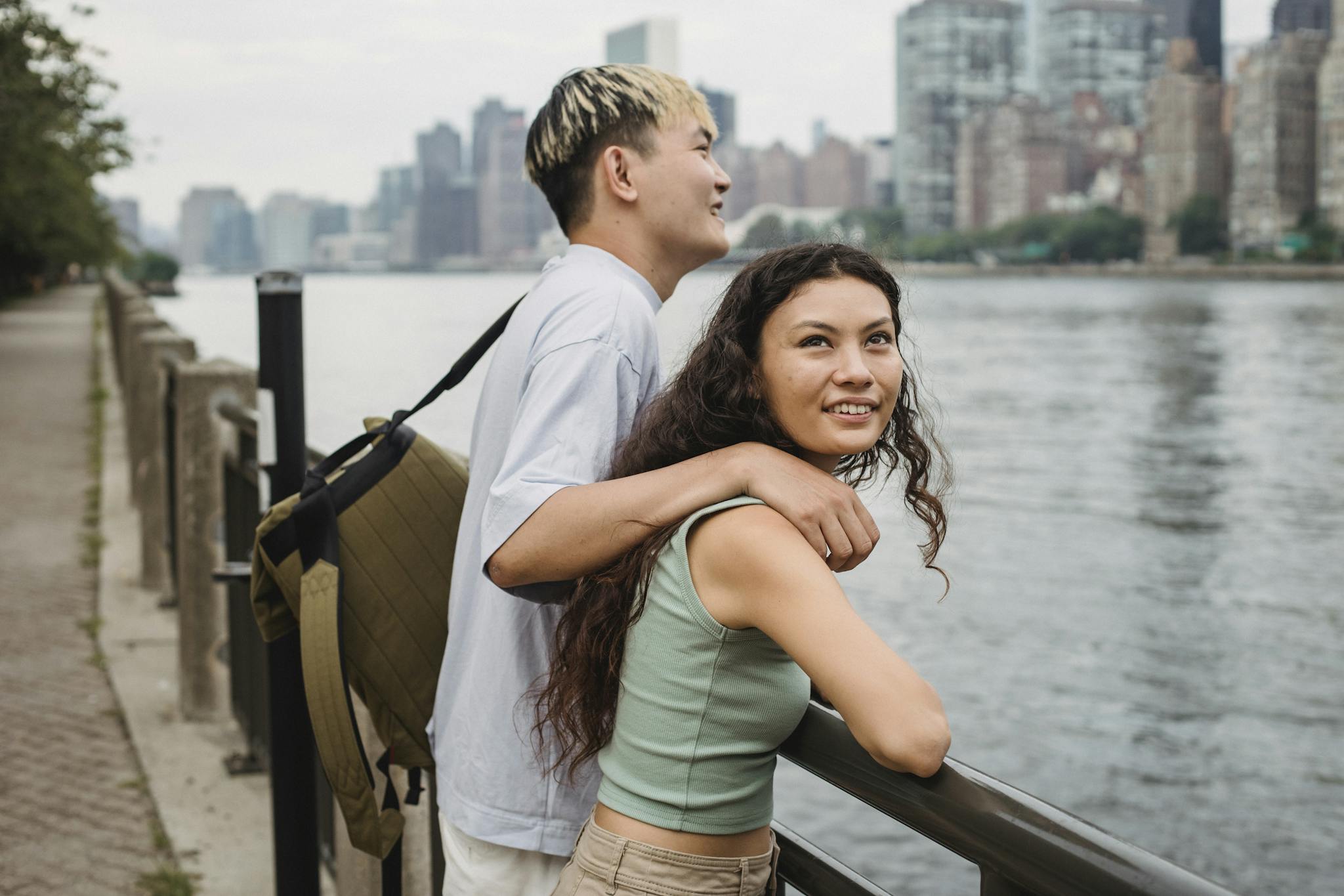 Side view of cheerful young diverse couple in casual clothes cuddling and smiling while standing on river embankment and sightseeing city