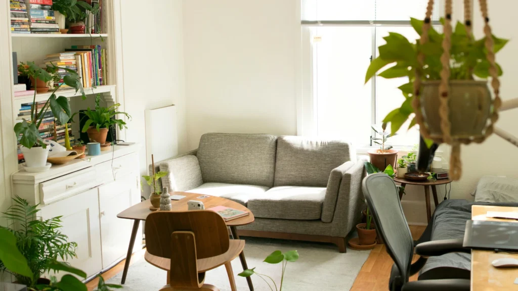 Ein kleines Zimmer, das in hellen Pastelltönen gehalten ist. Vorm Fester steht ein Sofa, davor ein Tisch mit einem Stuhl. An der linken Wand steht ein Regal, an der rechten ein Schreibtisch. Dazwischen sind verschiedene Grünpflanzen.