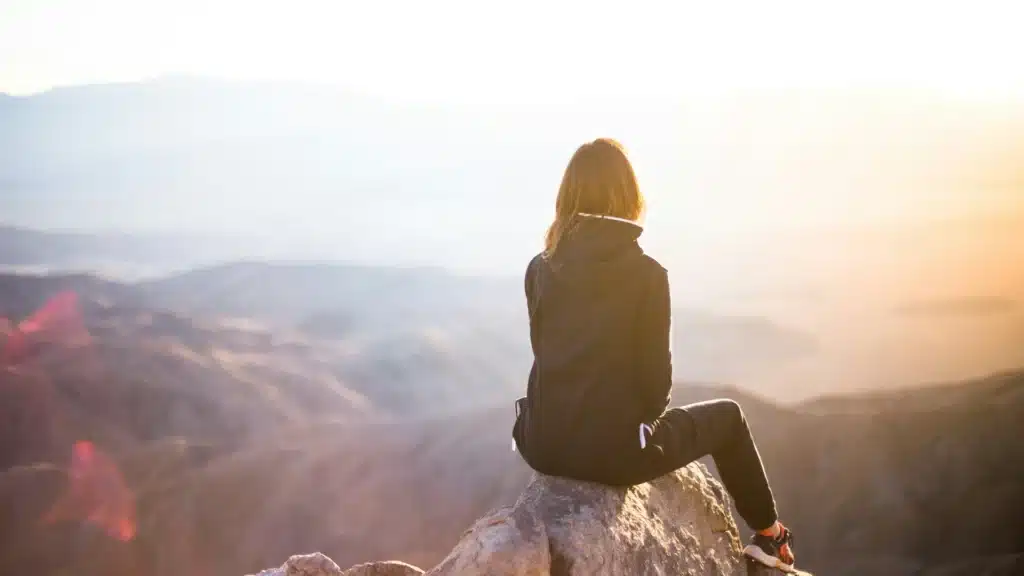 Eine Frau in Funnktionskleidung sitzt mit dem Rücken zur Kamera auf einem Gipfel. Ihr Blick schweift über weitere Berge im Hintergrund.