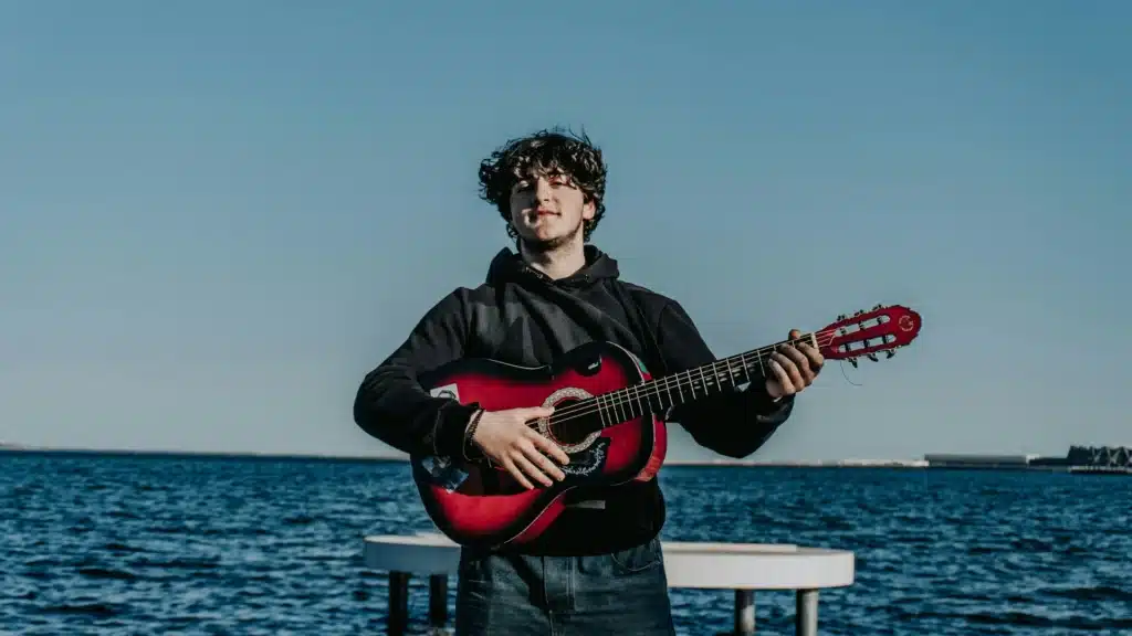 Ein junger Mann mit Gitarre steht am Ufer des Meerrs.