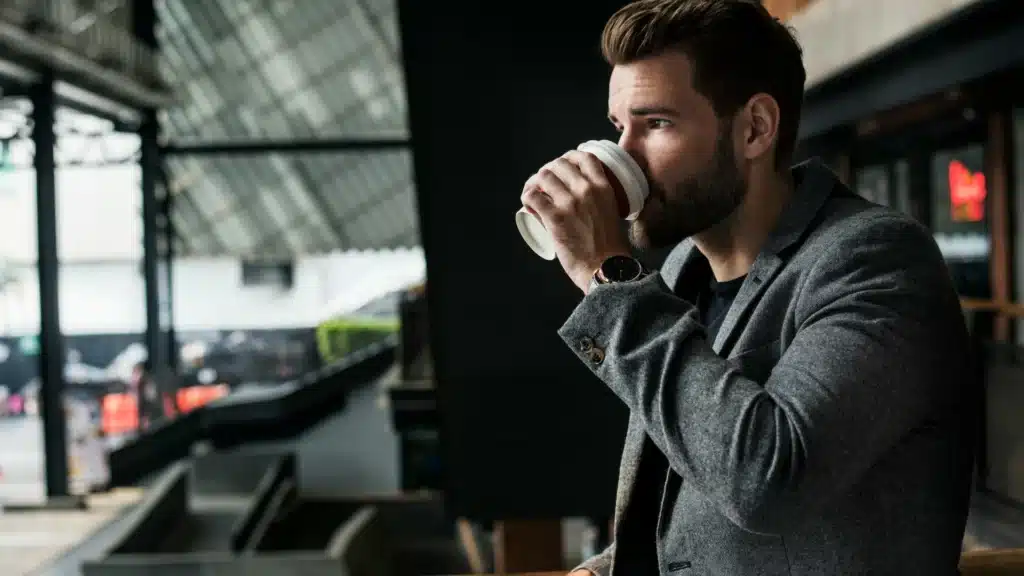 Ein junger Mann im Sakko steht an einer S-Bahn_Station. Er trinkt aus einem Kaffeebecher.