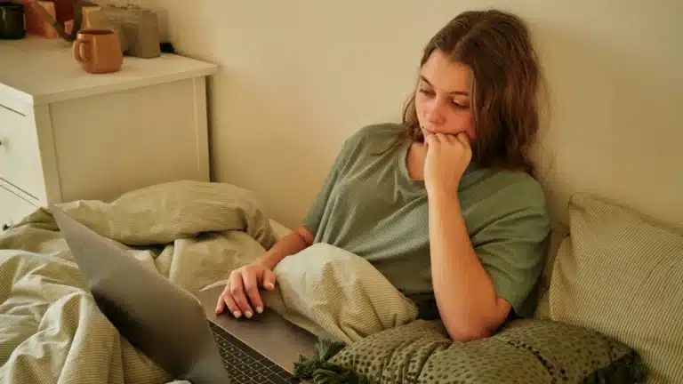 Eine junge Frau im olivgrünen T-Shirt liegt auf dem Rücken in ihrem Bett. Sie schaut auf dem Bildschirm eines Laptops, der vor ihr auf der Bettdecke liegt. Im Hintergrund sind weiße Schlafzimmermöbel zu sehen.