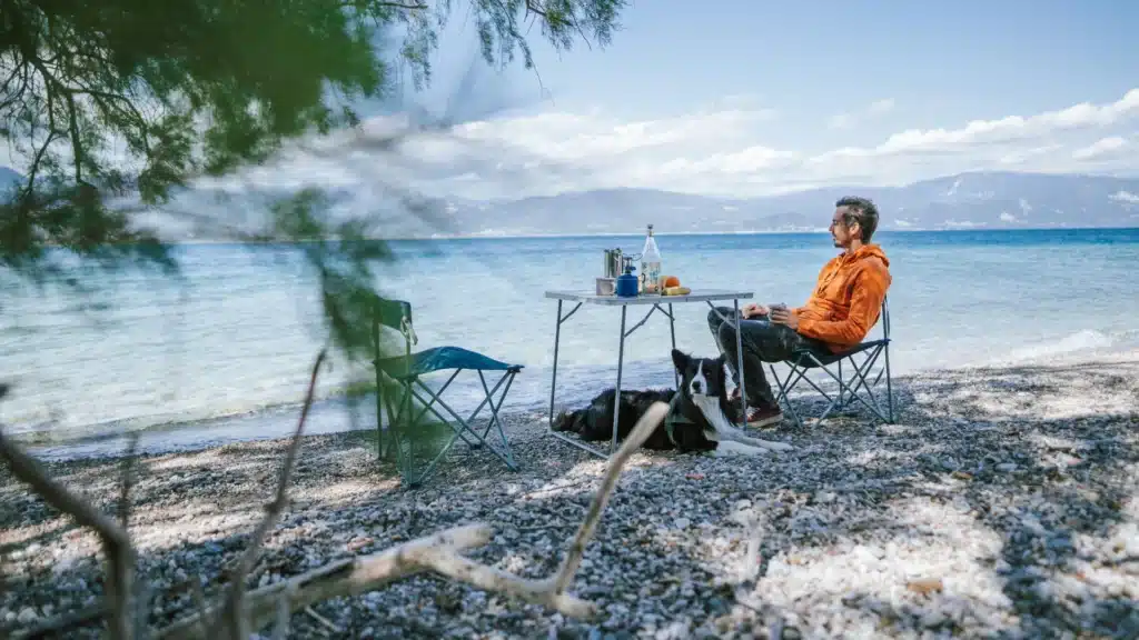 Ein Mann in orangenem Hoodie und Blue Jeans sitzt allein an einem gedeckten Campingtisch am steinigen Ufer eines Sees. Der Stuhl ihm gegenüber ist leer. Links ragt ein Baum im Bild, am anderen Ufer sind Berge zu erkennen.