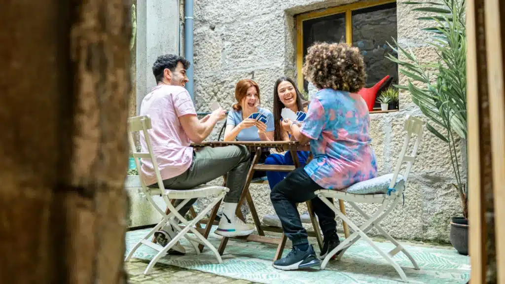 Ein Mann und drei Frauén sitzen in einem Innenhof auf Holzstühlen an einem Gartentisch. Sie lachen beim Kartenspiel. Im Hintergrund sind ein Fenster, eine Gießkanne und Grünpflanzen zu erkennen.