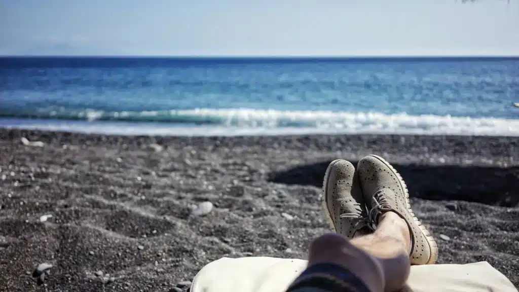 Ein Mann liegt auf einer Liege an einem Kiesstrand. Im Vordergrund sind seine grauen Lederschuhe zu sehen, dahinter das Meer und der Horizont.