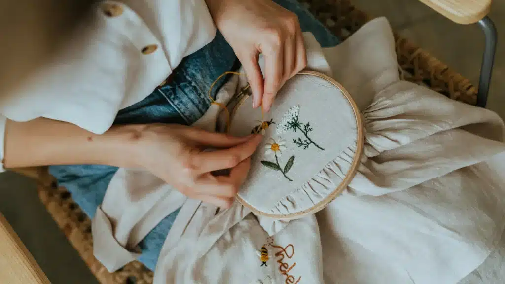 Eine Frau in weißer Bluse und Blues Jeans sitzt in einem Stuhl. Sie stickt ein Blumenmotiv.