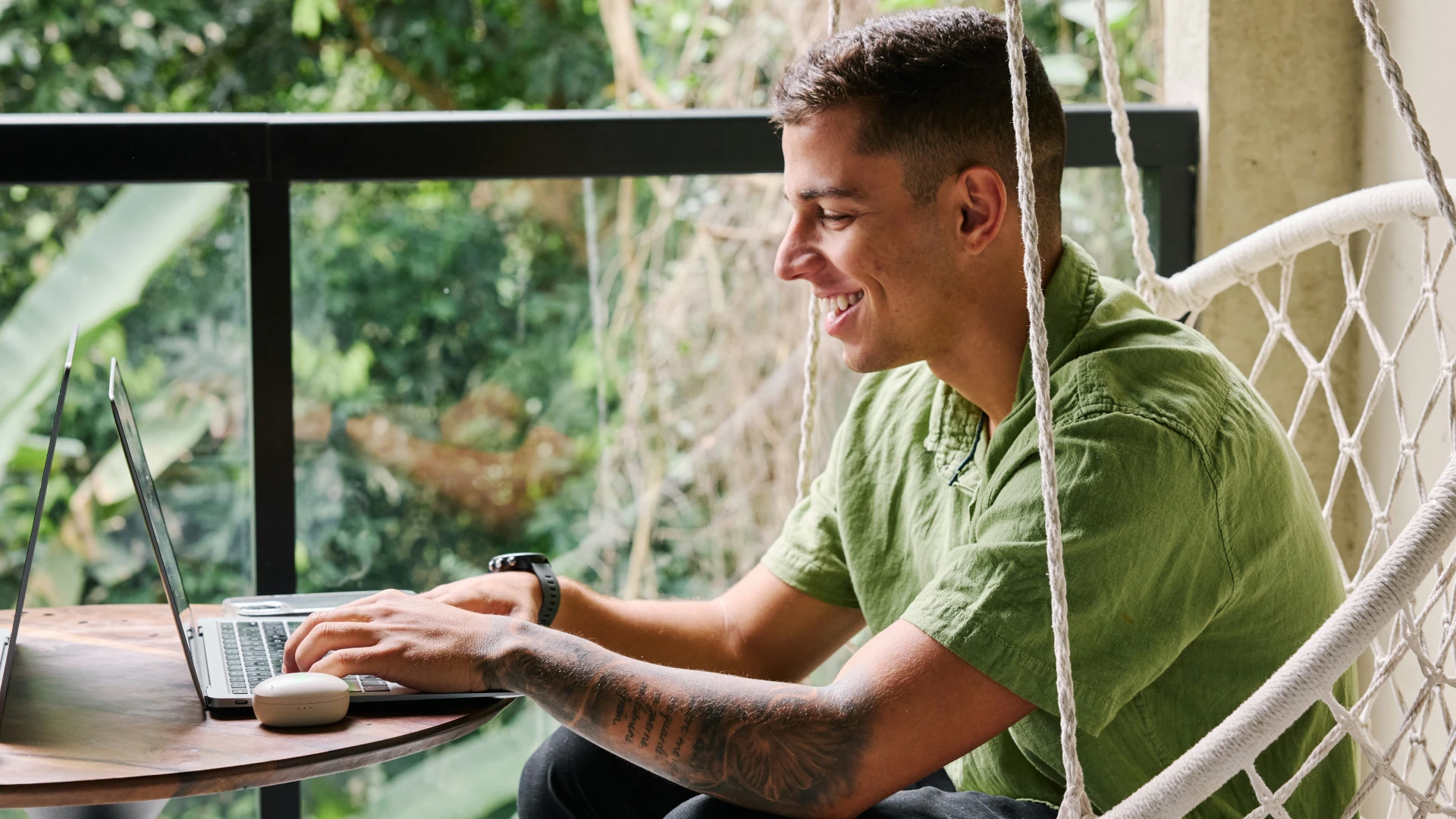 Ein braunhaariger Mann mit Tätoweirungen auf dem Arm sitzt in einem Hängesessel auf einem Balkon Er blickt lächenld auf den Bildsdchirm eines Laptops auf dem Tisch vor sich. Im Hintergrund ist üppiges Grün zu erkennen.
