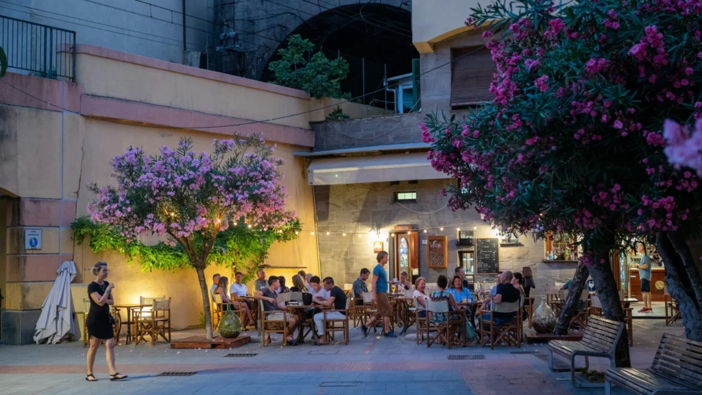 Auf dem Platz einer südlichen Stadt. Menschen sitzen unter einer Lichterkette an den Tischen eines Straßencafés. Bänke unter blühenden Bäumen laden zum Verweilen ein. Von links läuft eine einzelne schwarz gekleidete Frau durchs Bild.
