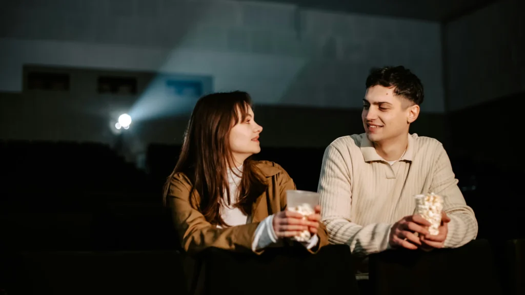 Eine Frau und ein Mann sitzen nebeneinander im Kino Beide werden durch ein Schlaglicht erhellt und halten je einen Becher mit Popcorn in ihren Händen.