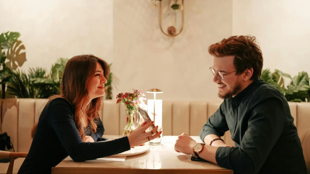 Eine Frau und ein Mann sitzen sich in einem Café mitt üppigem Blumenschmuck am Tisch gegenüber. Sie zeigt ihm das Display ihres Smartphones. Beide lächeln.