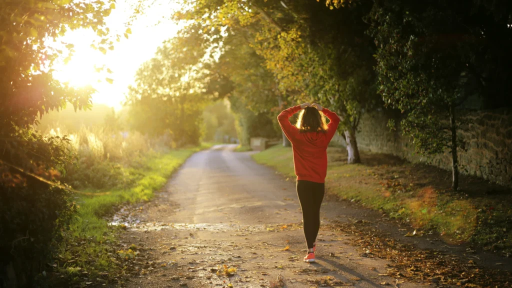 Eine Frau mit orangenem Hoodie und Schuhen sowie einer sxhwarzen Leggings läuft im herbstlich-nebligen Gegenlicht einen Weg zwischen Wald und Feld entlang. Sie hält ihre Hände über den Kopf.