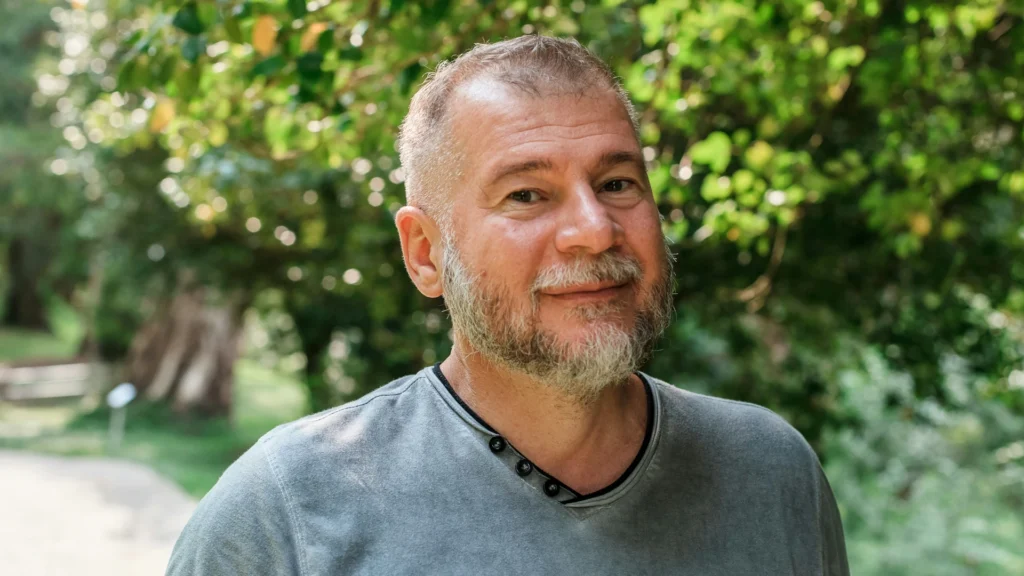 Ein Mann mit grau werdendem Bart und Haaren im grauen Shirt blickt mit leichtem Lächeln in die Kamera. Im Hintergrund sind Büsche zu erkennen.