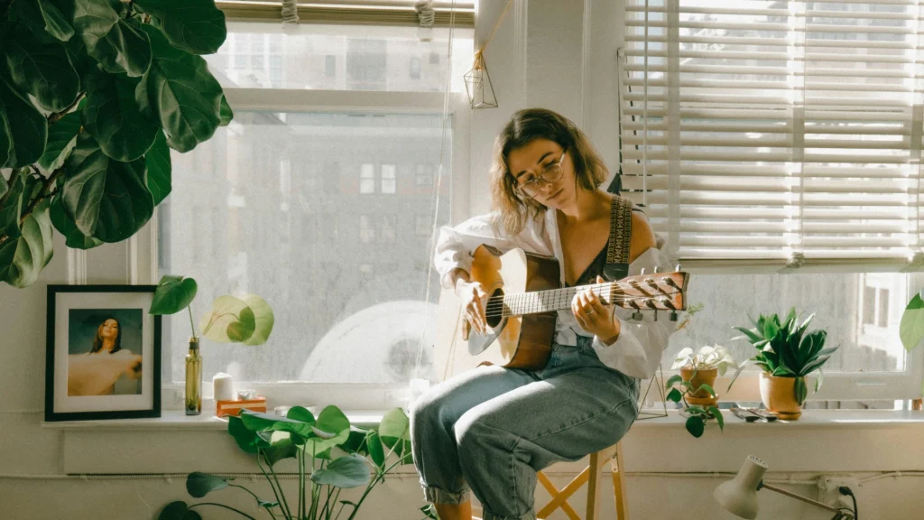 Eine Frau mit halblangen braunen Haaren und einer Brille sitzt auf einem Stuhl und soielt Gitarre Sie trägt eine weiße,weite Bluse und Blue Jeans. Auf der Fensterbank hinter ihr stehen Grünpflanzen, Fotos und Dekogegenstände. Durchs halbgeschlossene Roll fällt helles Sonnenlicht.