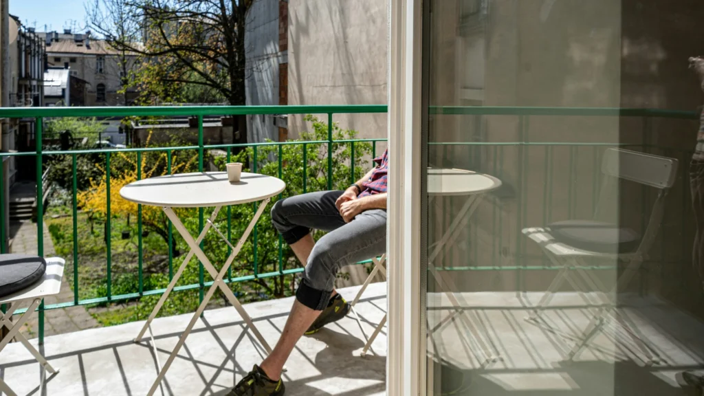 Eine Person sitzt in der Sonne auf einem Balkon. Von ihr sind nur die Beine zu sehen, die sich unter einen Tisch strecken. Auf diesem steht eine Kaffeetasse. Im Hintergrund sind Büsche, Bäume und Häuser zu sehen.
