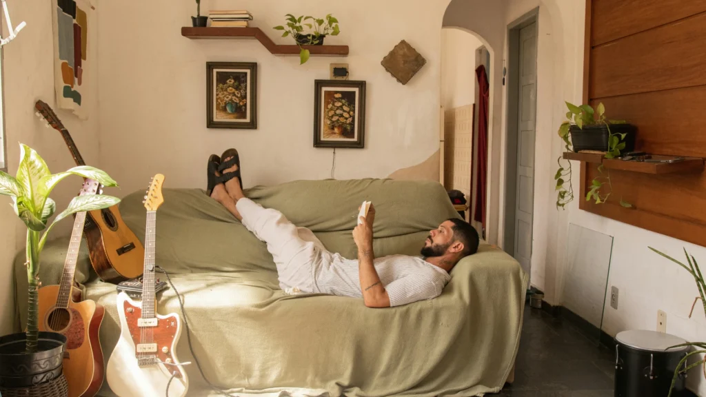Ein Mann liegt auf einem Sofa mit beigem Überwurf. Er hat die Füße in Sandaletten auf der Lehne und liest ein Buch. In Griffweite befinden sich Gitarren und Percussionsinstrumente. An den Wänden hängen Bilder und Regale mit Büchern und Grünpflanzen.