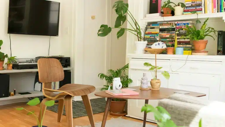 Ein Wohnzimmer. Auf einem braunen Laminat liegt ein weißer Wollteppich, darauf steht ein beiges Sofa, ein Topfmit einer Grünpflanze, eine brauner kleiner Tisch und ein brauner Holzstuhl. Rechts steht eine weiße Kommode mit Büchern und weitern Grünpflanzen, im Hintergrund befindte sich ein graues Sideboard, auf dem Plattenspieler und Verstärker stehen. Darüber hängt ein flacher Fernseher an der Wand.