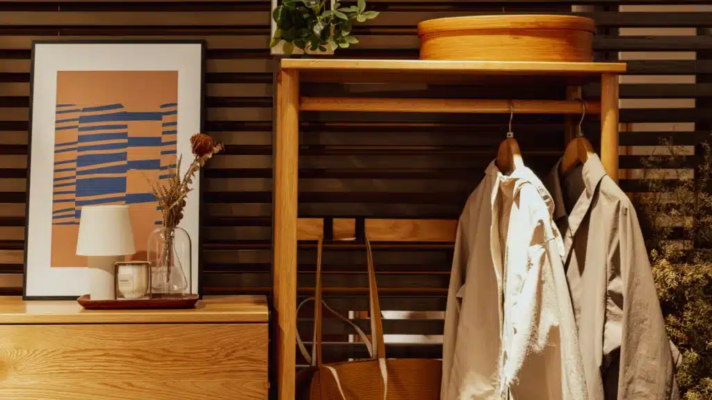 Eine hölzerne Garderobe. An der Wand befinden sich Akustikpaneele, davor rechts ein Garderobenständer mit Kleidungsstücken und einer Tasche, links eine Kommode, auf der ein Bild und verschiedene Deko-Gegenstände stehen.