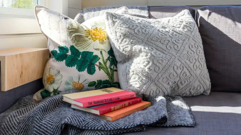 Auf einem blauen Sofa liegen links zwei Kissen, eiine Decke und drei Bücher. Ebenfalls von links fällt Sonnelicht durch ein Fenster.