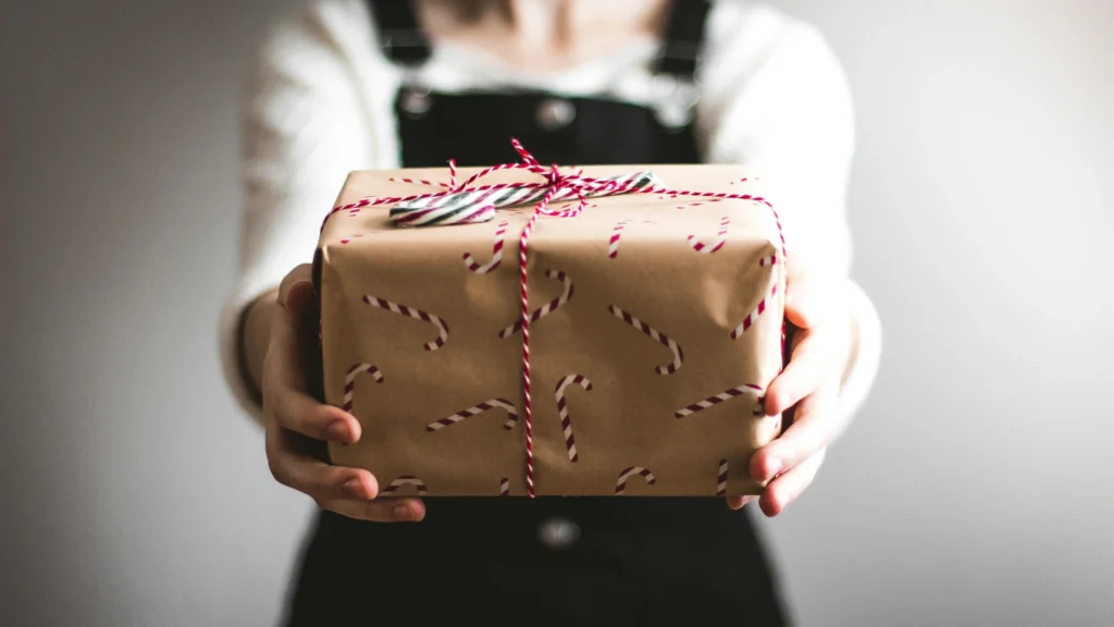 Eine Frau in weißem Pullover und dunkler Latzhose hält ein Geschenk in die Kamera- Es ist in braunes, mit Zucherstangen bedrucktes Papier verpackt und wird durch ein rot-weißes Band zusammengehalten.