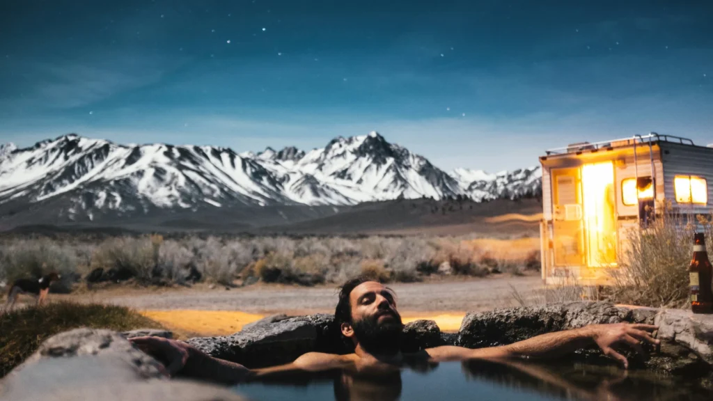 Ein Mann liegt mit ausgebreiteten Armen und geschlossenen Augen in einem Natursteinpool. Im Hintergrund sind eine erleuchtete Hütte und verschneite Berge zu sehen.