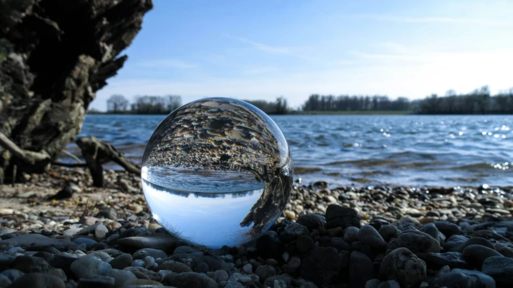 Auf einem Kiesstrand liegt eine Glaskugel, dahinter befindet siche eine Wasserfläche. Am gegenüberliegenden Ufer stehen Bäume hinter dem Schilfgürtel. In der Glaskugel spiegelt sich das Motiv so, dass es auf dem Kopf steht.