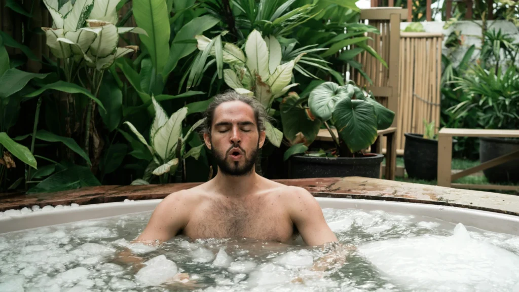 Ein braunhaariger, vollbärtiger Mann sitzt vor üppigen Grünpflanzen im Hintergrund in einer Badewanne. Auf der Wasseroberfläche treiben Eisstücke.