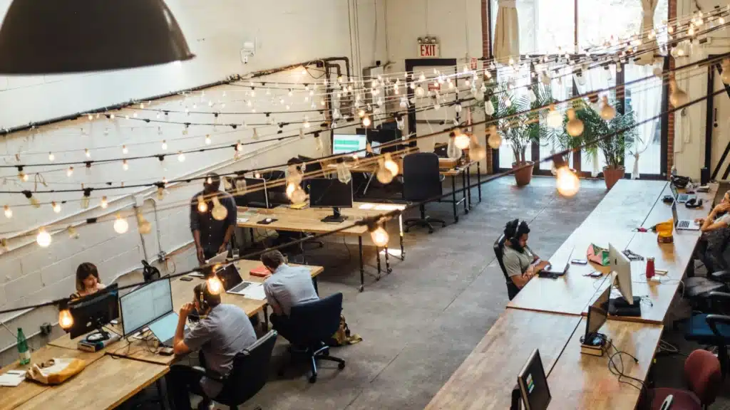 Ein Coworking-Space mit großen, fahrbahren Holztischen mit schwarzen Gestellen. Mehrere Menschen sitzen über den Raum verteilt an ihren Rechnern. Die unverputzten Wände sind weiß gestrichen, vor dem Fenster im Hintergrund stehen große Grünpflanzen in Terracottatöpfen. Über den Arbeitsplätzen hängen Lichteketten mit nackten weißen Glühlampen..
