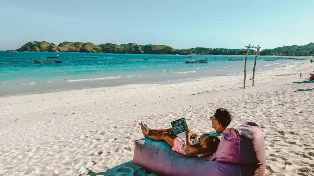 Ein Mann liegt auf einer aufblasbaren Liege an einem Sandstarand. Er blickt über einen Laptop, der auf seinen Beinen steht, hinaus aufs Meer. Weiter rechts am Strand sind eine Schaukel, ein Hund und weitere Menschen. In der Dünung liegen Fischerboote.