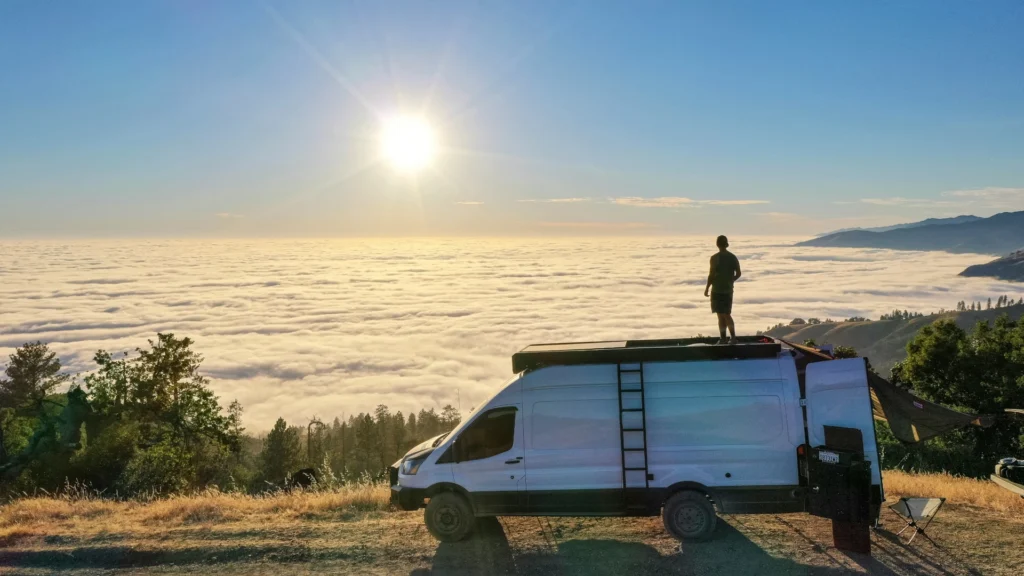 Ein Mann steht mit dem Rücken zur Kamera auf dem Dach eines Transporters, der am Rand eines Feldwegs geparkt ist. Er blickt über bewaldete Hänge hinab auf ein Wolkenmeer, über dem die Sonne steht.