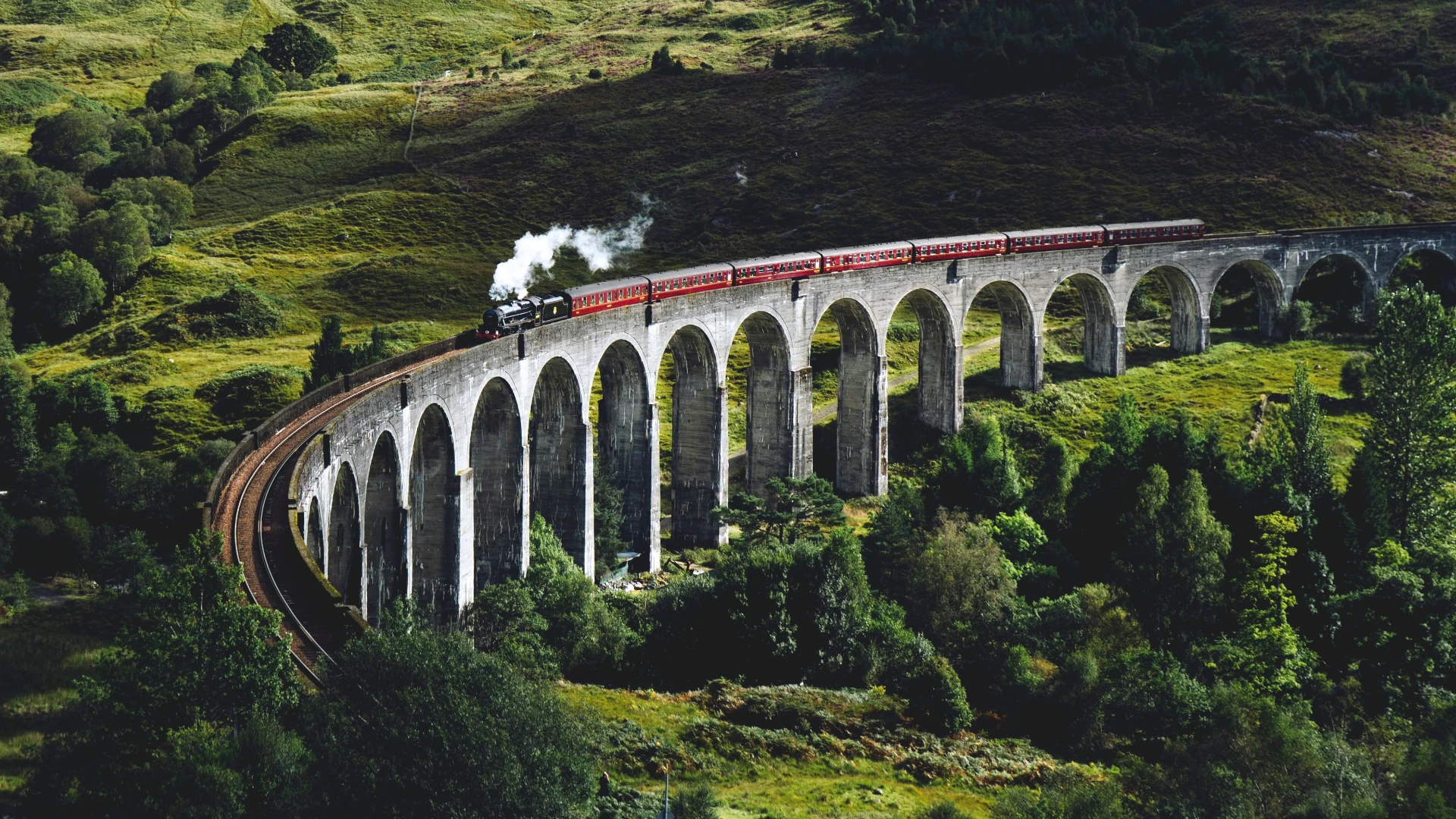 Ein mit Dampflokomotive bespannter Zug aus roten Wagen befährt einen gebogenen Steinviadukt in einer Hügellandschaft.