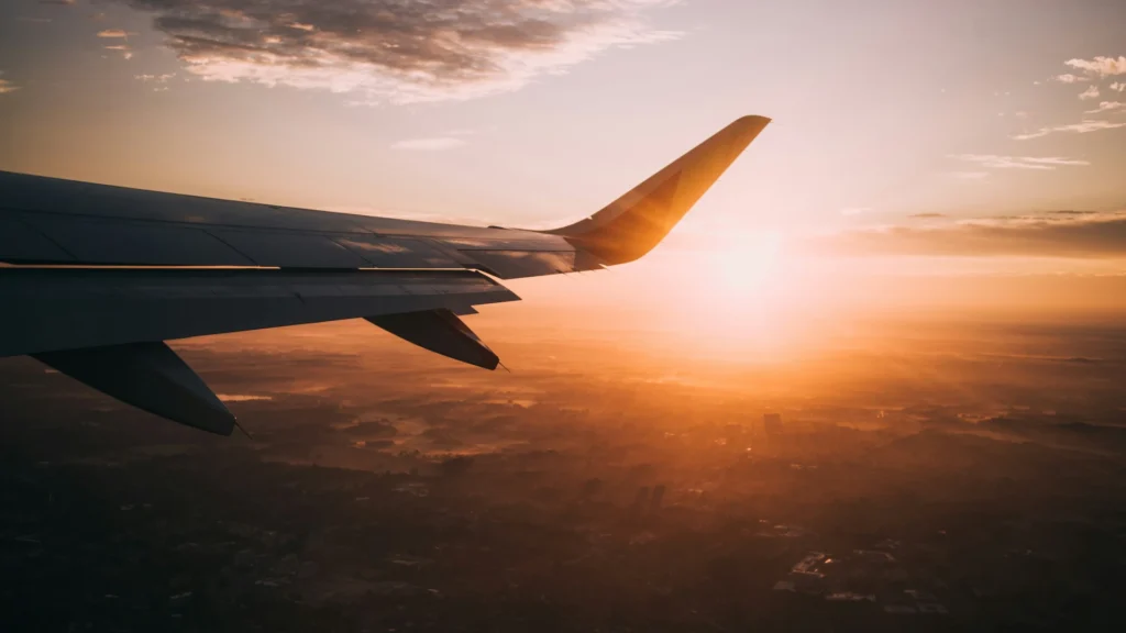 Blick aus einem Flugzeugfenster. Neben der rechten Tragfläche geht die Sonne unter. Darunter ist schemenhaft eine Landschaft zu erkennen.