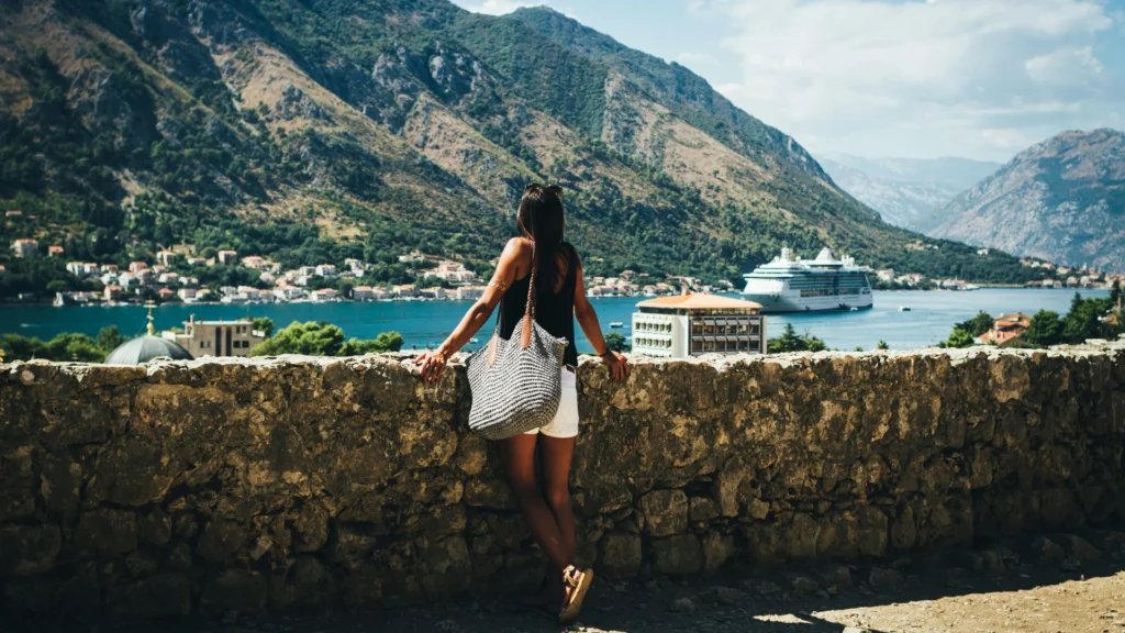 Eine Frau in sommerlicher Kleidung und Umhängetasche lehnt mit dem Rücken zur Kamera an einer Steinmauer. Sie blickt auf die Häuser an einem Fjord, auf dem ein Kreuzfahrtschiff schwimmt.