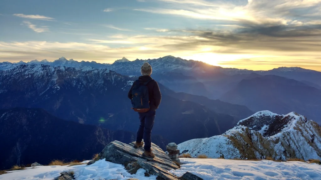 Ein Mensch in Winterkleidung und Rucksack steht mit derm Rücken zur Kamera auf einem Stein, der aus einem Schneefeld ragt. Er blickt in einen wolkenverhangenen Sonnenuntergang über einer Bergkette.