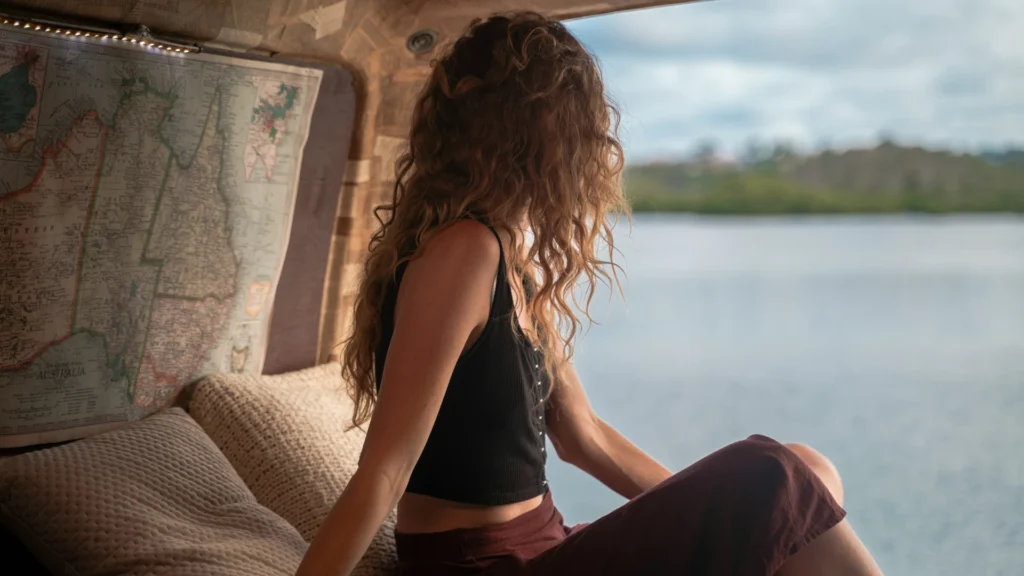 Eine Frau in einem schwarzen Trägerhemd und einer braunen Caprihose sitzt auf dem Bett eines Campers. Hinter ihr an der Wand hängt eine Weltkarte. Sie blickt mit abgewandtem Kopf auf einen See.