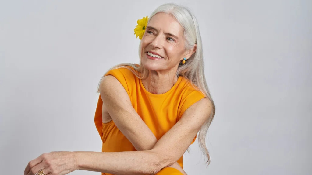 Eine ältere Frau mit langen weißen Haaren schaut lächelnd schräg nach oben an der Kamera vorbei. Sie trägt ein orangefarbenes, ärmelloses Kleid und hat eine gelbe Blüte im Haar.