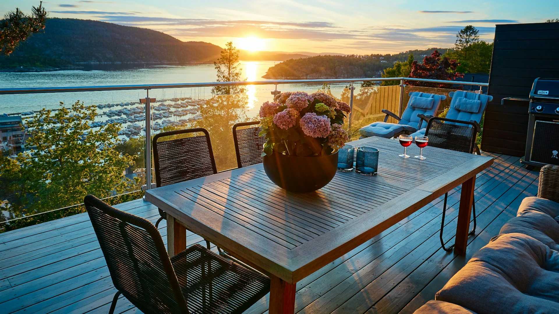 Ein geräumiger Balkon am Abend. Am Tisch stehen mehrere Stühle, auf ihm Weingläser, Teelichtständer und ein Blumenstrauß. Rechts hinten steht ein Grill. Der Blick geht durch weiter auf einen See und Berge, hinter denen die Sonne untergeht.