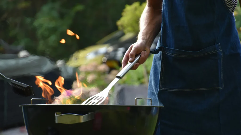 Ein Mensch mit Schürze steht mit Wender in der rechten Hand an einem Kugelgrill, aus dem Flammen lodern.