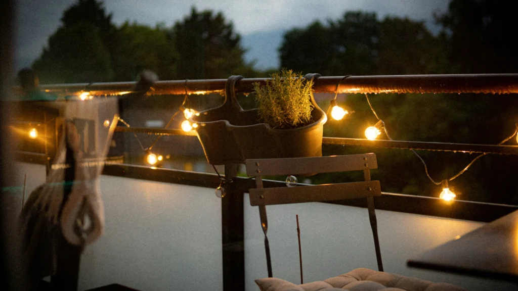 Ein Balkon bei Dämmerung. Ein Stuhl steht an einem Tisch. An der Brüstung hängt ein Blumenkasten mit Kräutern sowie eine Lichterkette, die die Szene mild beleuchtet.