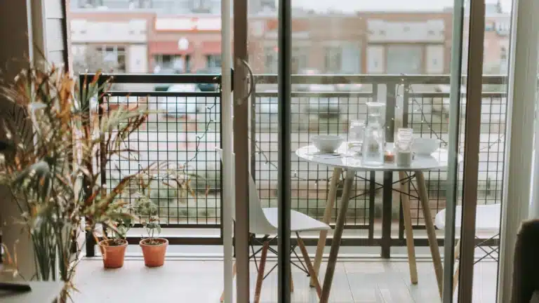 Auf einem Balkon steht ein Tisch mit zwei Stühlen. Auf dem Tisch ist für ein Frühstück zu zweit gedeckt. Links stehen drei Pflanzen in Töpfen auf dem Boden. Im Hintergrund ist unscharf eine Industriehalle zu erkennen.