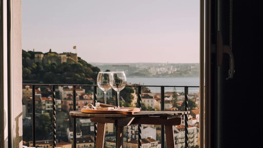 Zwei Weingläser stehen auf einem kleinen Balkontisch. Der Blick gleitet über eine Stadt. Links steht eine Burg auf einem Hügel, rechts spannt siche ine Brücke über den Fluss.