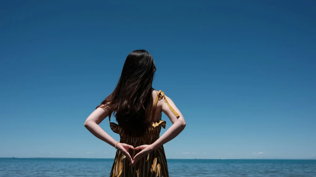 Ein Frau mit dunklen Haaren und champagnerfarbenen Kleid steht mit dem Rücken zur Kamera am Meer. Sie hat die Hände hinter ihrem Rücken verschränkt.
