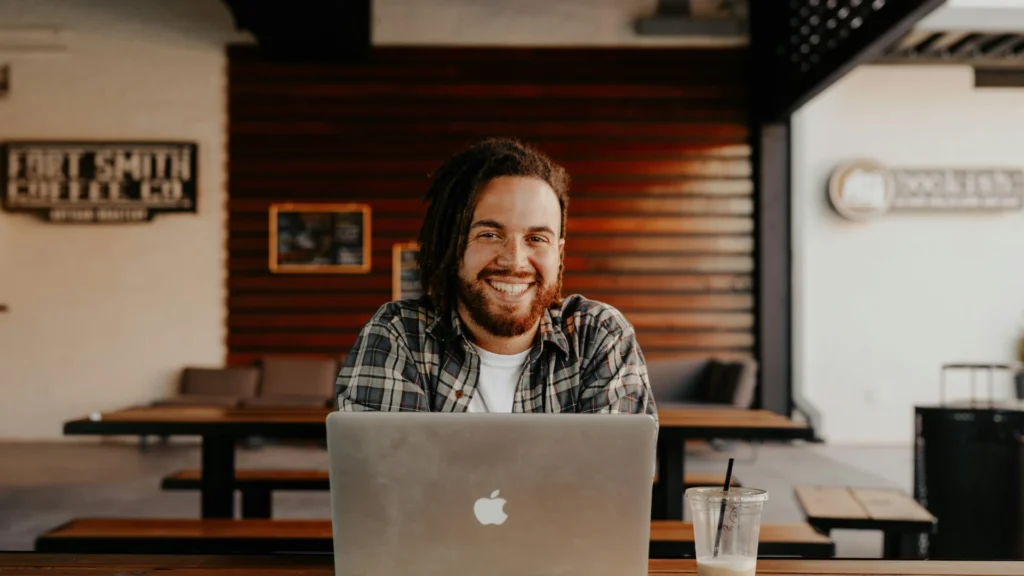 Ein Mann mit braunen Haaren und Vollbart sitzt vor seinem Laptop in einem Coworking Space. Er lächelt in die Kamera.