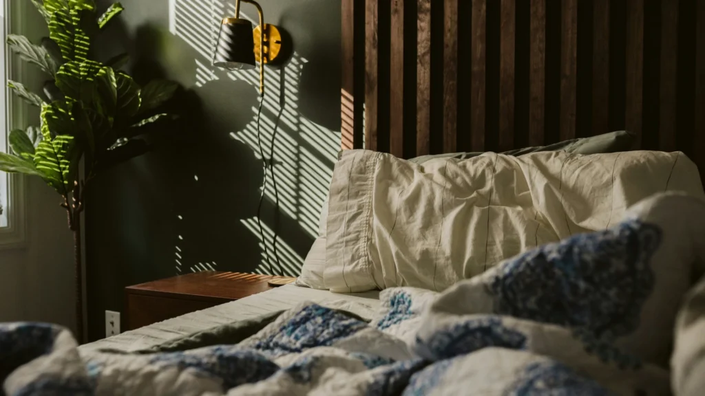 Ein Bett mit aufgeworfener Decke und mehreren Kissen. An der Wand dahinter ein Akustikpanel, daneben eine Leselampe, darunter ein hölzerner Nachttisch, in der Ecke eine Grünplanze. Von links fällt mildes Licht durch die Jalousien.