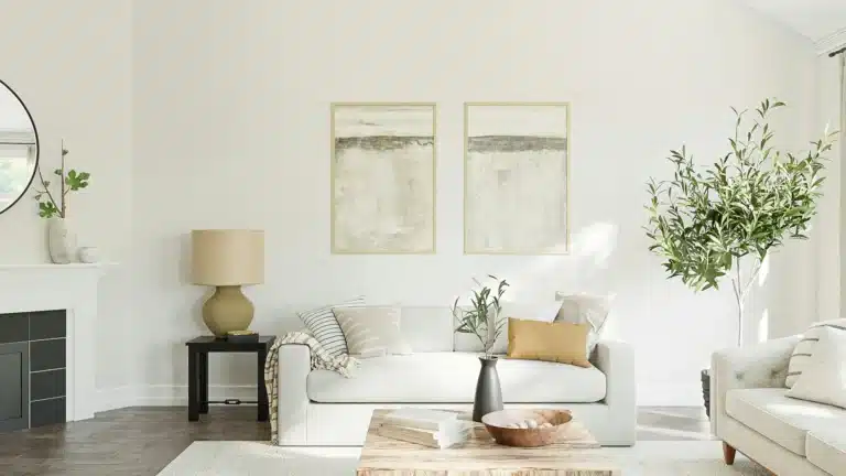 Ein Wohnzimmer mit zwei beigen Sofas, die über Eck stehen, Vor ihnen liegt ein ebenfalls beiger Teppich. An der Wand hinten hängen zwei abstrakte Fotos. Links ist ein schwarzer Beistelltisch mit einer Stehlampe, daneben ein Kamin.