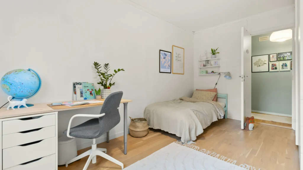 Eine Art Wohnheimzimmer. ÜBer dem Bett in der Ecke hängen Bilder und ein keines Bücherregal mit Leselampe. Daneben steht ein weißer Schreibtisch mit ebenfalls weißem Bürostuhl. Auf dem Tisch steht ein Globus, davpr liegt ein cremefarbener Teppich. Ein paar Pflanzen und Dekogegnstände sollen eine wohnliche Atmosphäre schaffen.