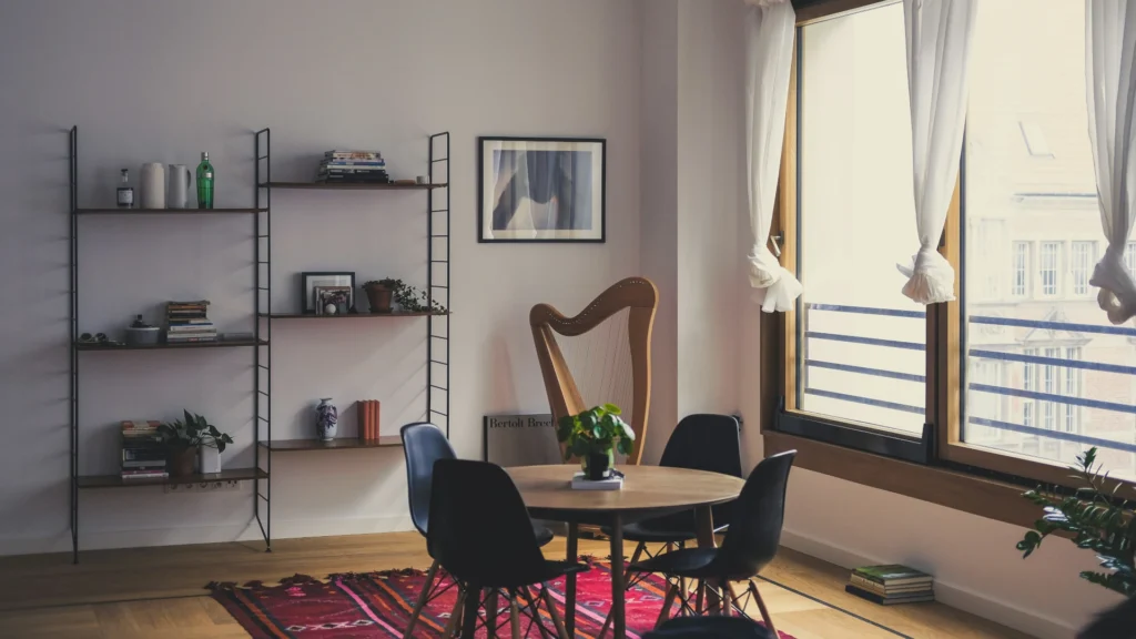 Ein sparsam möbliertes Wohnzimmer. Auf einem roten Teppich steht ein runder Tisch mit vier Stühlen. An der Wand dahinter hängt ein schlichtes Regal mit wenigen Büchern und Deko-Gegenständen, daneben lehnt eine Harfe an der Wand. Vor dem Fenster rechts hängen weiße, geknotete Gardinen.