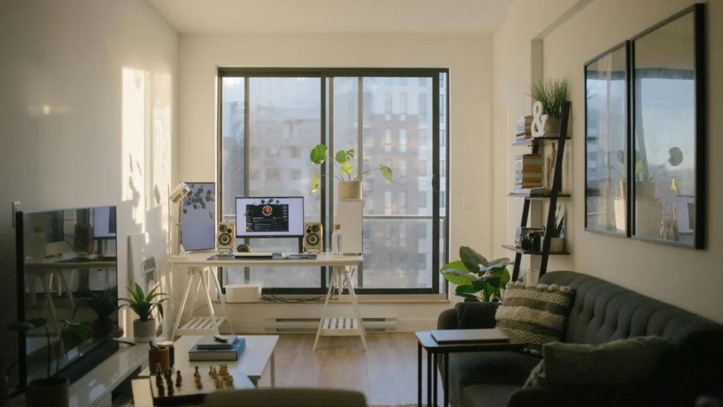 Ein Wohnzimmer. An der linken Wand hängt ein Flachbildschirm, davor steht ein Beistelltisch mit Büchern. An der linken Wand hängen zwei Bilder, davor steht ein dunkles Sofa, dahinter ein Regal mit Büchern. Den Platz vor dem bodentiefen Fenster im Hintergrund nimmt eine Platte auf zwei Böcken ein, die Tastatur und Bildschirm trägt. Einige Grünpflanzen ergänzen das Bild.