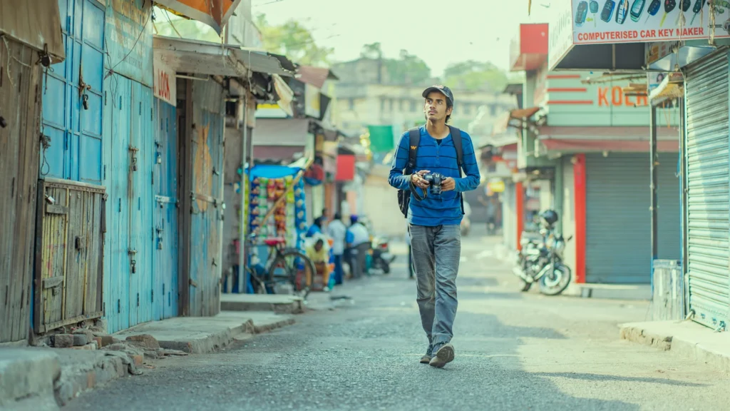 Ein Mann mit Basecap, blau-schwazem Shirt, Blue Jeans und schwarzem Rucksack geht durch die Straße einer Stadt vermutlich in Asien. Er hält eine Kamera in seinen Händen-