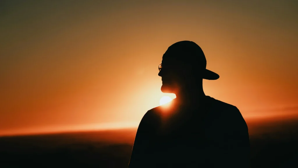 Ein Mann mit Basecap und Brille steht mi dem Rücken zur Kamera vor einem Sonnenuntergang.