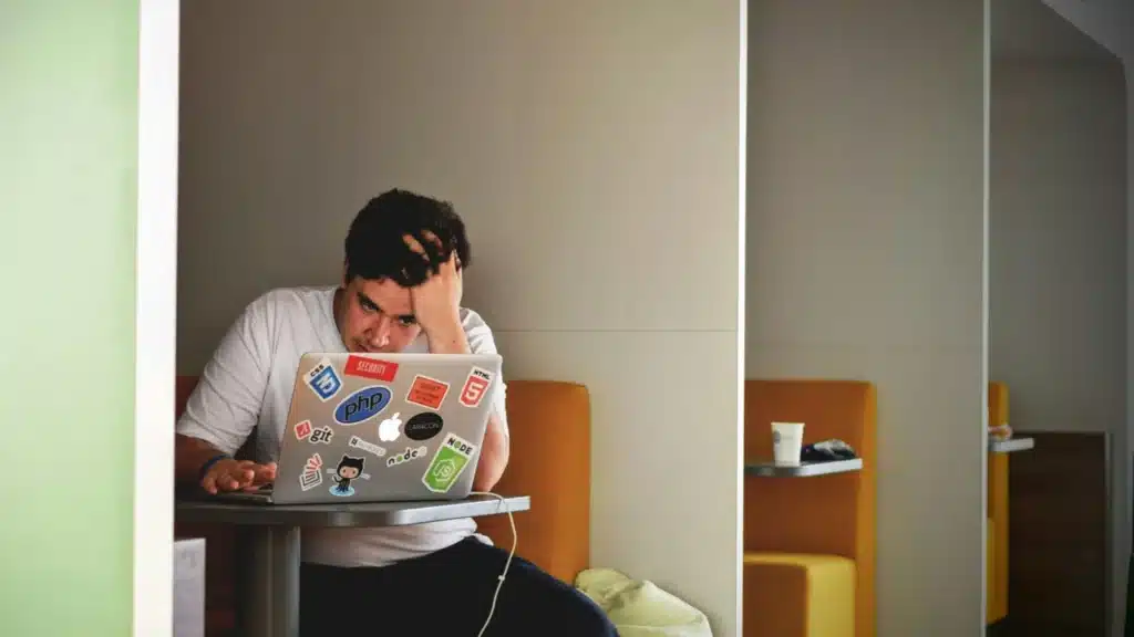 Ein Mann sitzt ein einer Nische eines Cafés. Er stützt den Kopf auf seine linke Hand und blickt konsterneiert auf den Bilschirms des Laptops, der vor ihm auf dem Tisch steht.